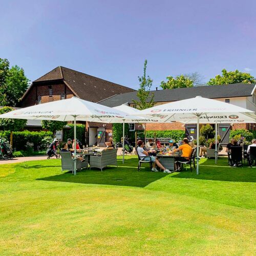 Menschen sitzen an Tischen unter großen Sonnenschirmen auf einer grünen Terrasse vor Gebäuden an einem sonnigen Tag