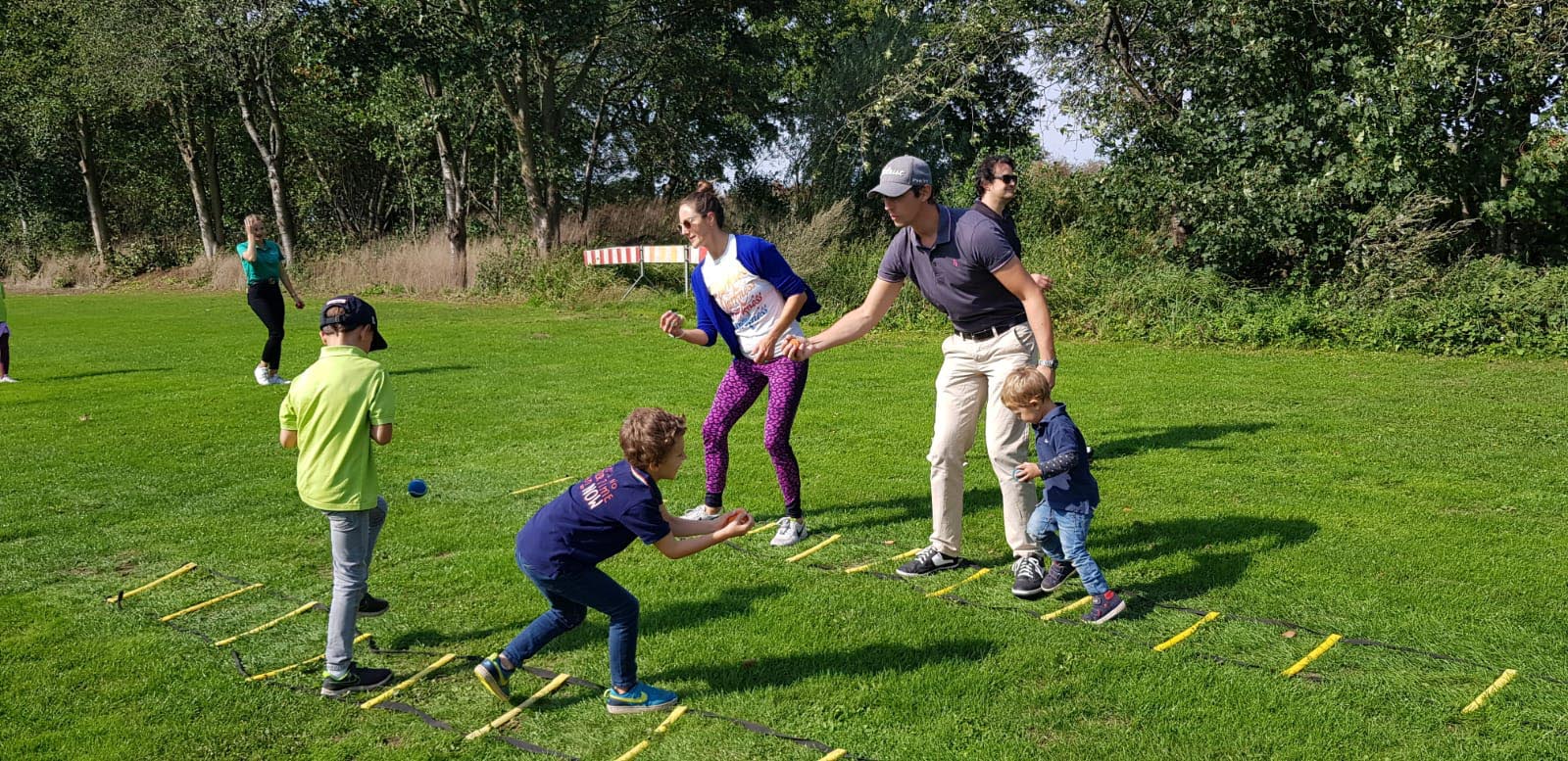 Mehrere Kinder und Erwachsene spielen gemeinsam im Freien auf einer Wiese mit Koordinationsleitern und Bällen